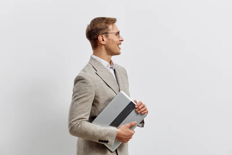 Professional young man in formal suit holding a folder, smiling confidently on plain light gray background. Business, success, professional, confident, 30s, corporate, office style.