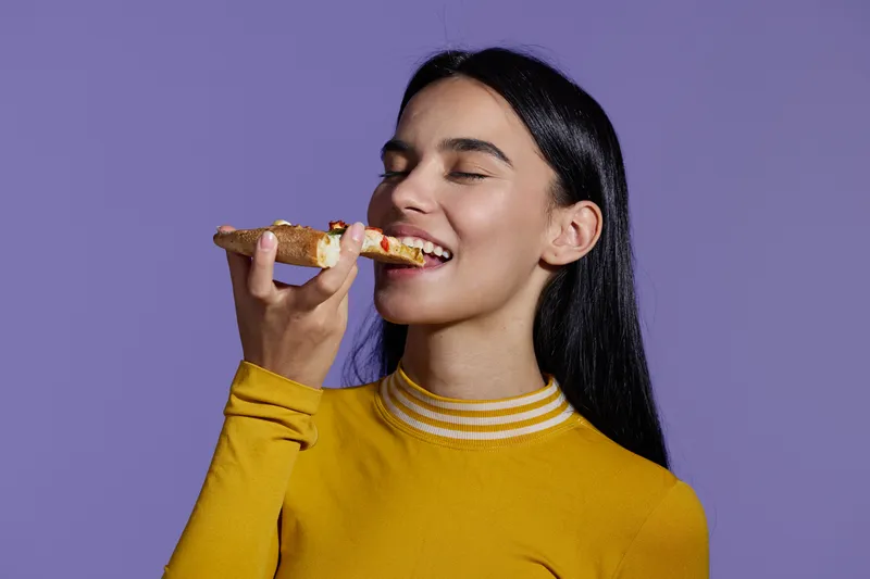 woman sport fitness enjoying a healthy lifestyle, smiling young woman with long black hair eating food against plain purple background, bright casual outfit