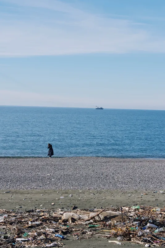 Solitary figure walking along a pebble beach as calm sea stretches to the horizon, litter and debris in the foreground creating a contrast between nature and pollution.