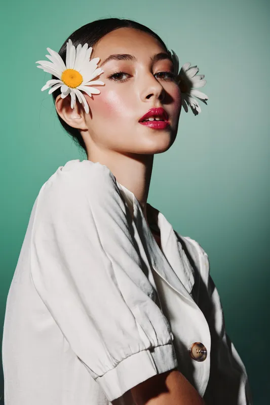 Young woman with floral accessories in hair wears a white jacket and shiny makeup, posing confidently against a pale green background in a creative beauty fashion studio shot.