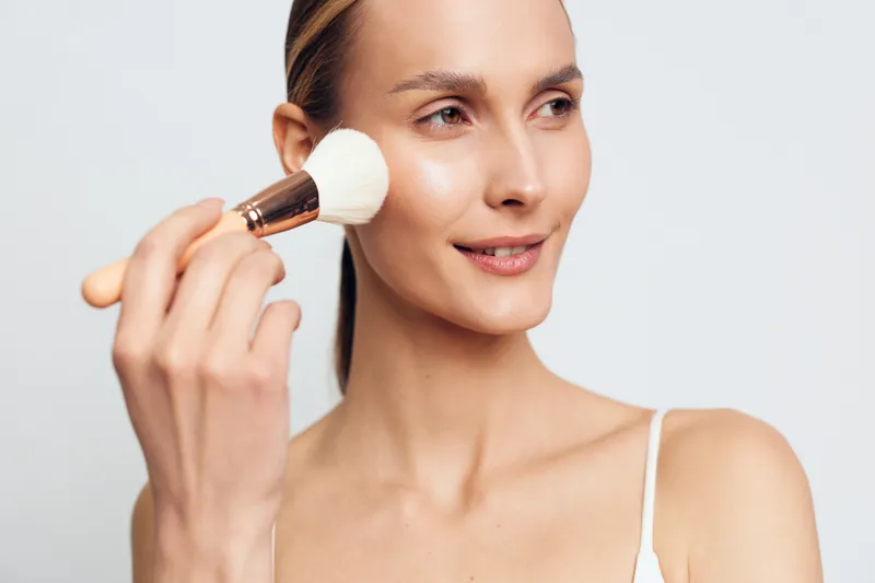 Young woman applies makeup with powder brush on face smiling gently looking sideways isolated on white background in beauty skincare concept.