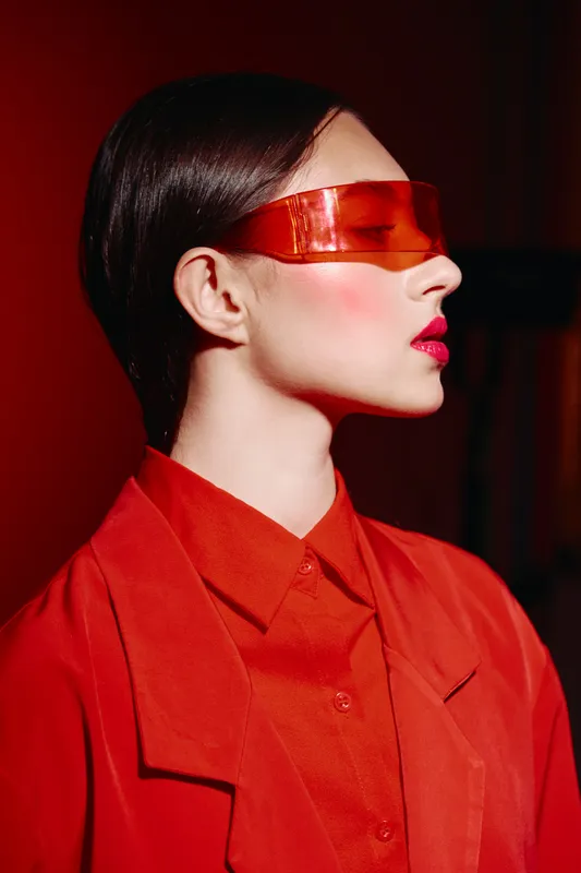 Female model in upscale red clothing wears glossy futuristic blindfold over eyes, posing in contrast between crimson shade and skin tone in studio. Concept of fashion creativity and beauty.