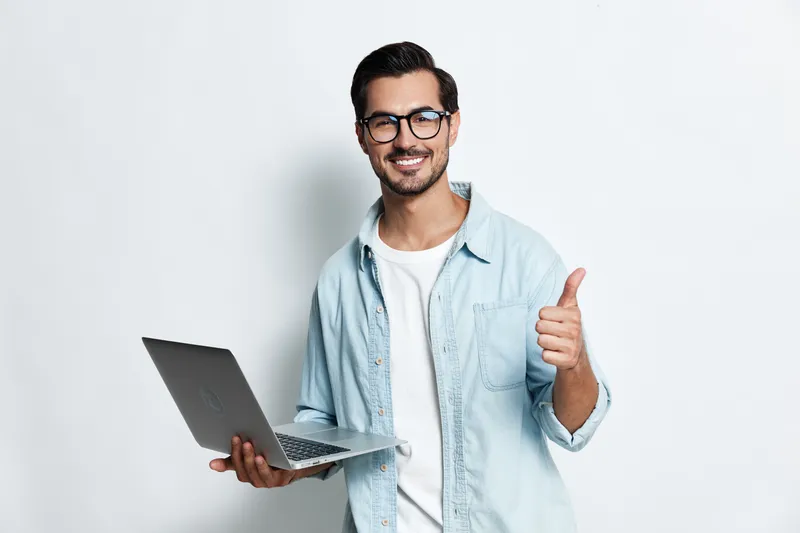 Happy smiling young man holds laptop in one hand and gives thumbs up with the other, showcasing excitement and success against a plain white background.
