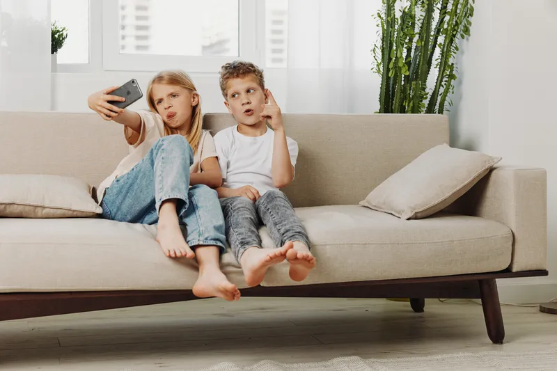Two kids sitting barefoot on beige sofa making selfie with smartphone in bright living room. Casual children enjoy playful moments at home with relaxed expressions.