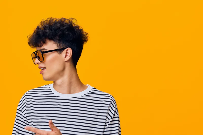 young man with curly hair wearing striped t-shirt and sunglasses looking away against vibrant yellow background, casual style and cheerful mood