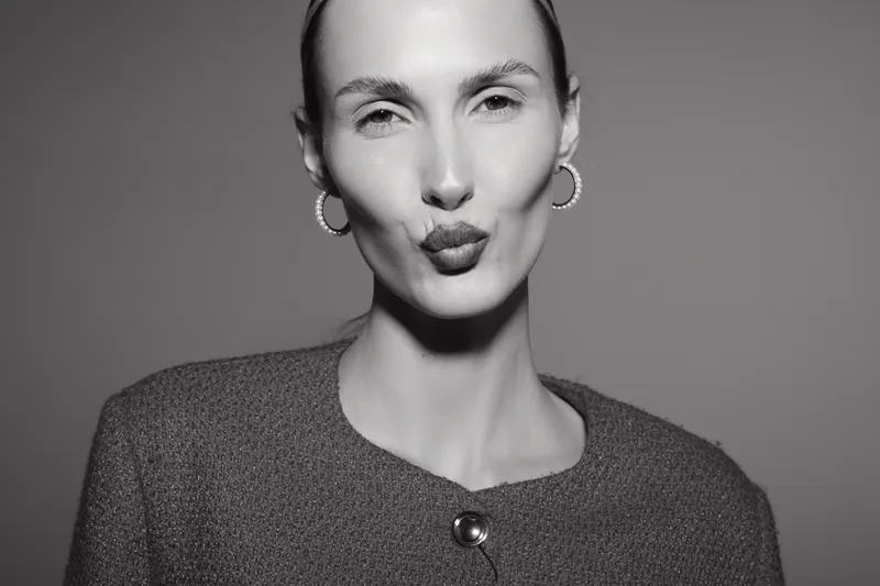 White woman with pale skin and slicked back hair makes a kissy face in studio black and white portrait.