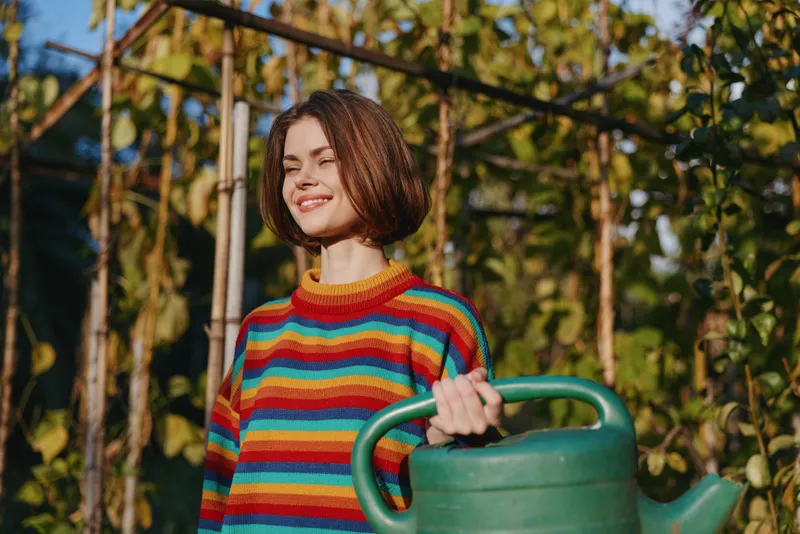woman holding a watering can in a garden wearing a rainbow sweater, smile with eyes closed in sunlight among green plants and trellis, outdoor gardening and summer care