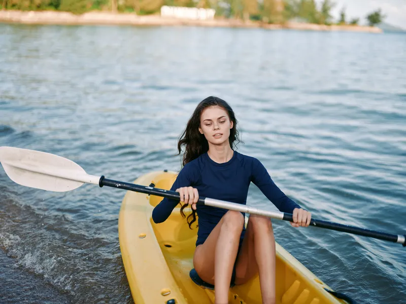 A young woman paddles a yellow kayak on calm water, wearing a dark blue long sleeve top, focused expression as she rows with a double paddle, sunlight glinting on the ripples.