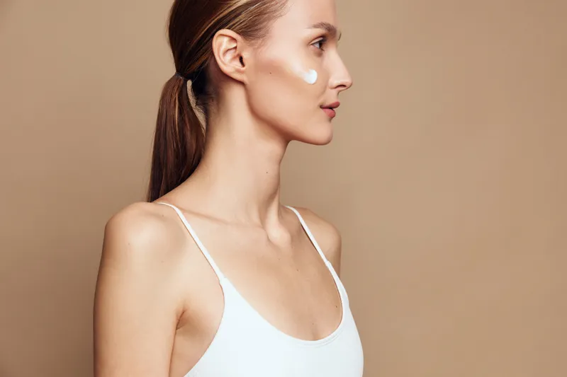 Young woman with clear skin applying white face cream on cheek, side profile on beige background. Concept of skincare, beauty routine, and natural wellness. Soft lighting highlights smooth skin.