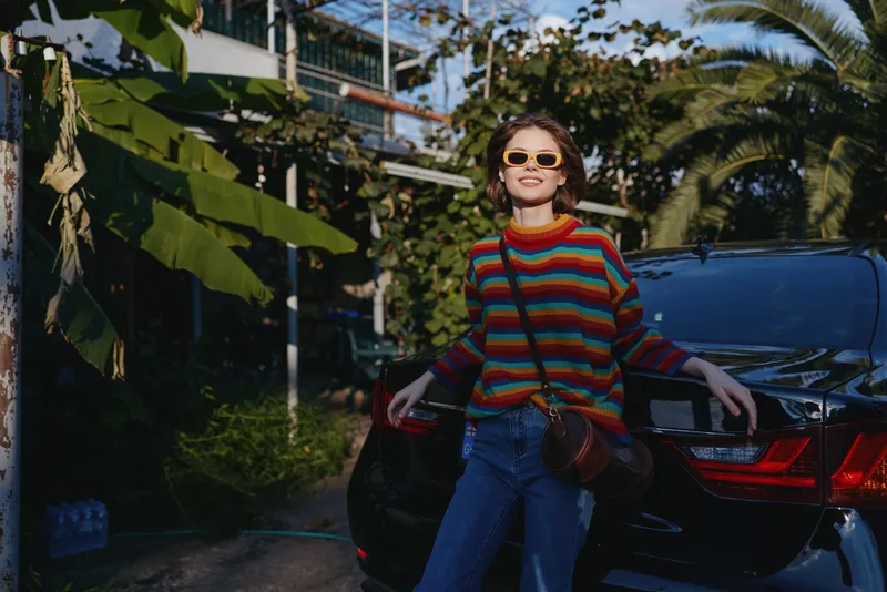 Woman striped sweater sunglasses leaning car tropical smiling casual outfit with crossbody bag and denim jeans, sunlit travel lifestyle portrait in a relaxed urban tropical street scene.