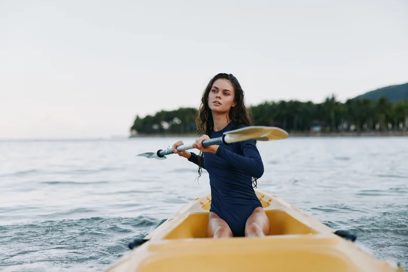 Young woman kayaking on calm ocean waters with green island background in warm natural light during daytime.