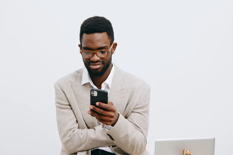 Modern young man with glasses using smartphone in an office setting, dressed in casual blazer, focused on screen, clean white background, technology lifestyle concept, professional appearance, communi