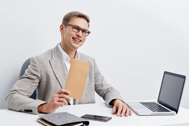 Smiling professional man in smart casual attire holding a notebook while sitting at a desk with a laptop, tablet, and smartphone on a plain white background. Business, technology, workspace concept.