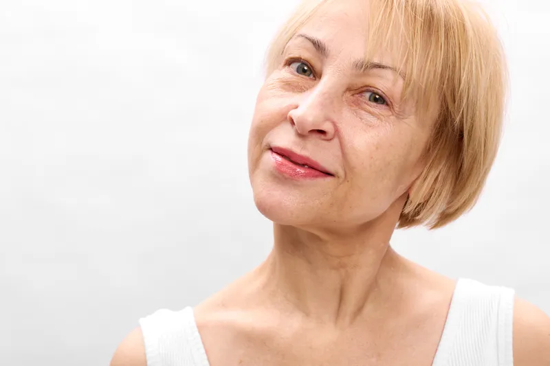 Senior blonde woman closeup portrait, gentle smile, soft studio lighting, white background, calm mature face, focused eyes, skincare health wellbeing care
