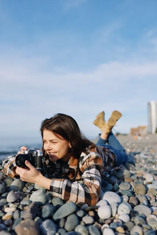 Woman photographer lying on pebbles at the beach, capturing coastal scenery with a camera, enjoying outdoor leisure and nature in a sunny seaside moment