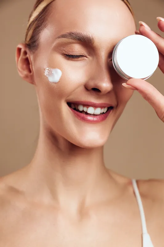 Woman skincare face beauty cosmetic cream application closeup, smiling with eyes closed, hand holding cosmetic jar, gentle soft lighting, calm home setting, smooth skin, beige background