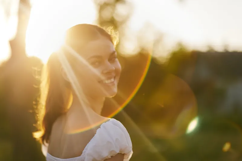 Smiling young woman in a sunlit outdoor setting, dressed in a light summer dress, radiating joy and warmth during the golden hour