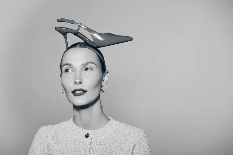 Portrait of a fair skinned Caucasian woman with slicked back hair wearing a high heel shoe on her head, posing against a plain grey background in black and white.