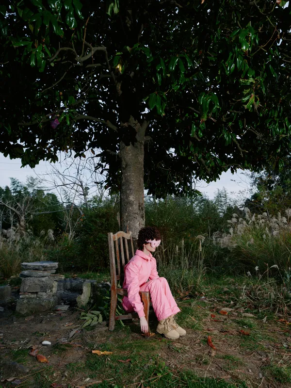 Girl sitting on a wooden chair under a large tree in a calm outdoor setting, wearing a pink dress and boots, surrounded by grass and scattered leaves.
