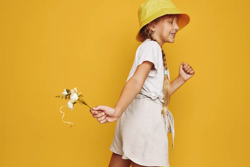 Young girl in beige dress and yellow hat holds cotton flowers walking smiling on yellow background. Studio shot, playful and cheerful mood.