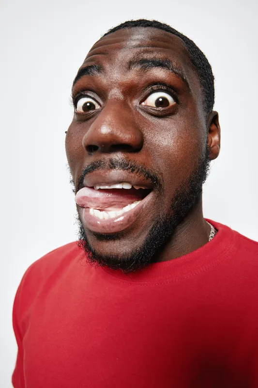 African American man with dark skin making funny face with wide eyes and tongue out wearing red shirt on white background. Closeup portrait with playful expression and beard.