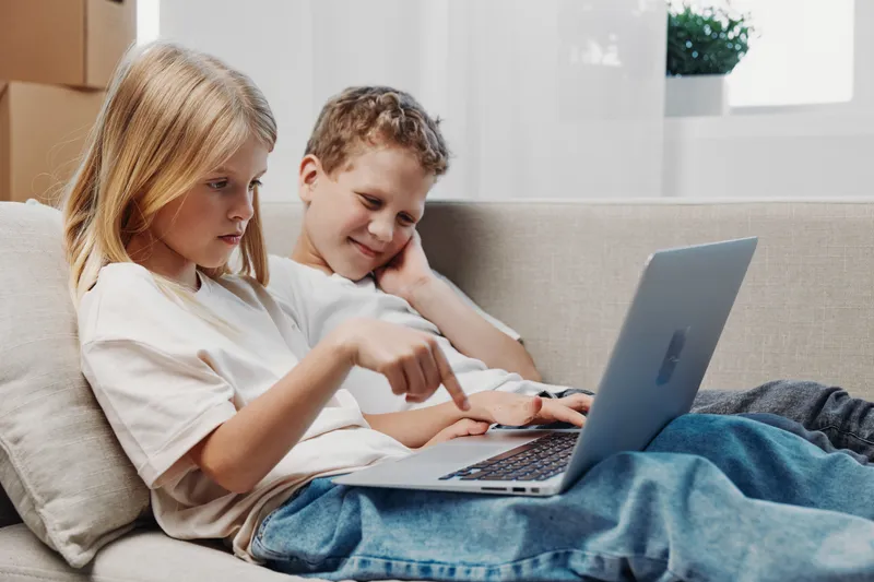 Two children sitting on a couch using laptop together, focused and engaged. Cozy home environment with natural light, concept of learning, technology, and childhood interaction.