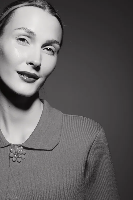 Portrait of a Caucasian woman with pale skin wearing a textured sweater with a brooch, looking confidently at camera in a black and white studio setting. Beauty and fashion concept.