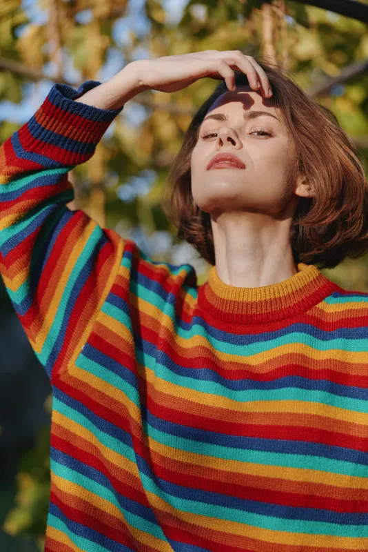 Young woman in a striped sweater with short brown hair in a sunlit outdoor portrait, 25-35 years old, confident pose shielding eyes, casual outfit and colorful knitwear in garden setting.