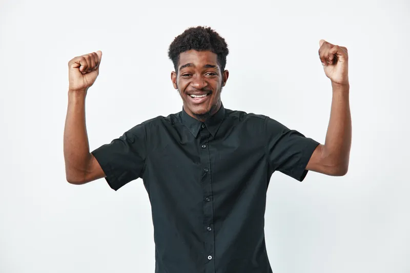 Young black man with dark skin wearing black shirt smiling and raising fists in celebration isolated on white background. Happy African ethnicity male expressing success and joy.