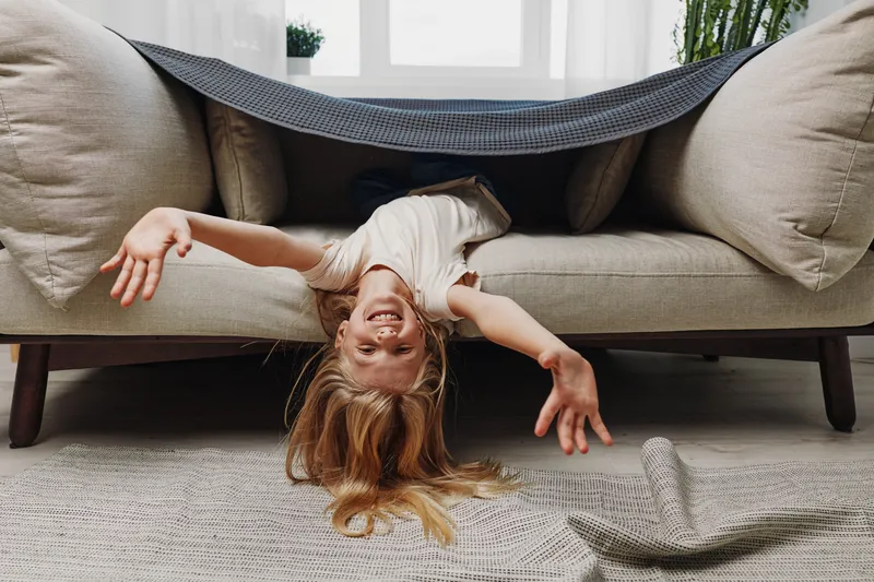 Playful child with long hair lies upside down on beige sofa under a blanket, smiling and reaching out with arms in a cozy living room. Concept for childhood joy and home comfort.