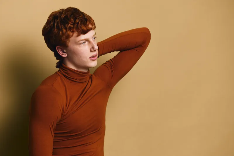 Young man with red hair wearing brown turtleneck posing with hand behind head against beige background looking thoughtful in studio portrait photography.