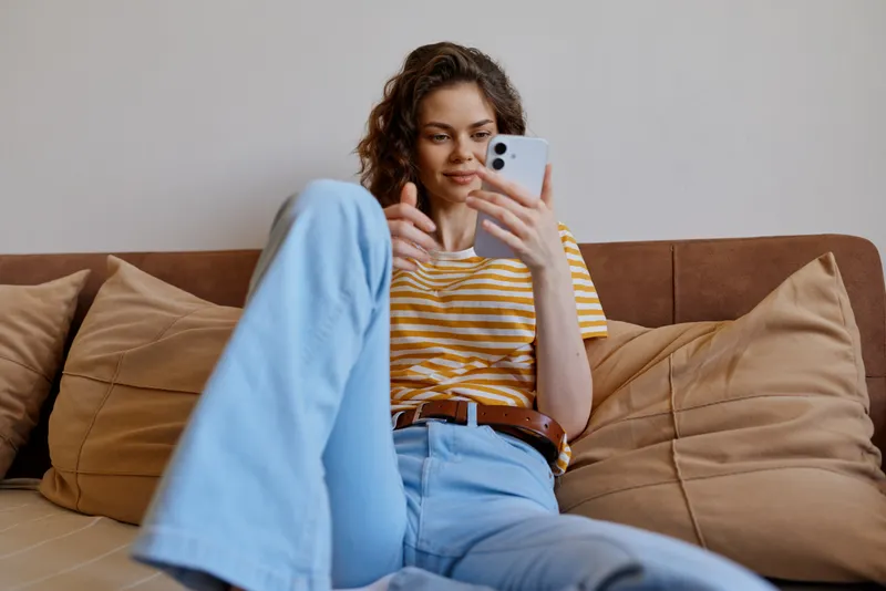 A young woman with curly hair sits on a sofa at home, browsing her smartphone, looking focused and relaxed, capturing a candid moment of everyday life.
