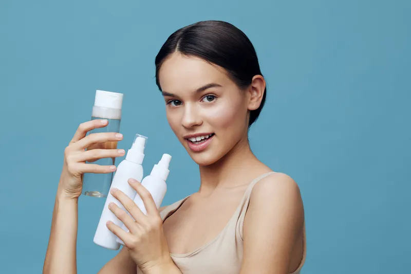 Young woman holding skincare products, smiling with a light blue background, showcasing healthy skin and beauty concept, ideal for cosmetics marketing