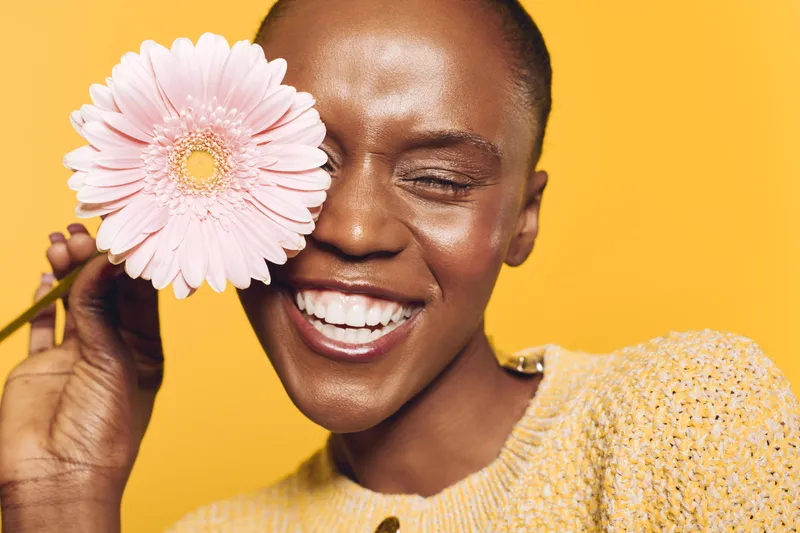Happy African woman with dark skin smiles broadly holding a pink flower near her face. Bright yellow background and warm lighting create a joyful and vibrant mood of natural beauty.