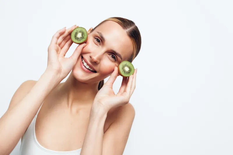 A smiling young woman holds two halves of fresh kiwi fruit near her face against a white background. Concept for healthy lifestyle, natural beauty, and skincare.