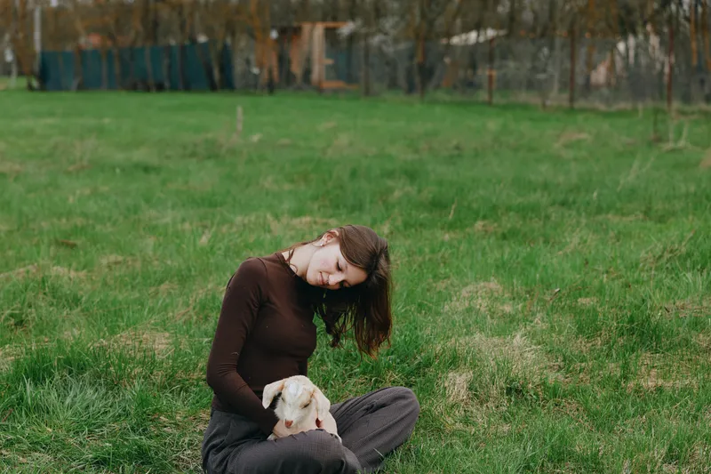 A woman in a brown sweater sits on the grass, cradling a gentle goat kid in her arms in a wide field. A calm lifestyle moment illustrating connection with animals, nature, and outdoor living.
