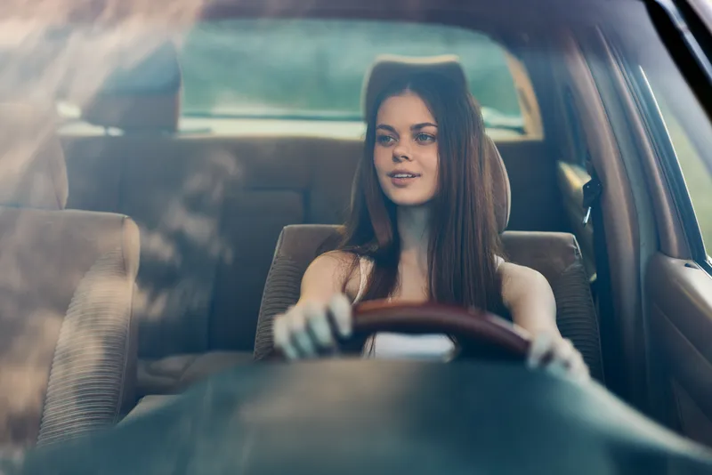 Young woman driving a car with a joyful smile, wearing casual attire, in a natural setting characterized by soft lighting and a warm color palette.