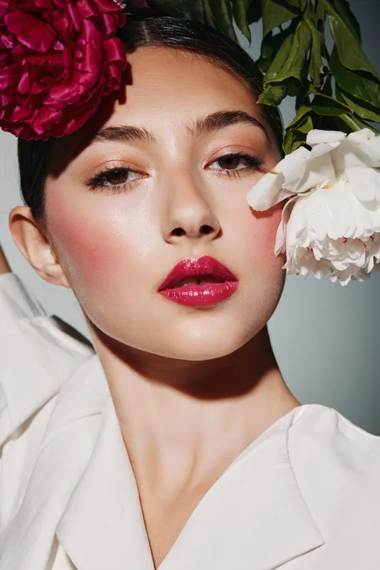 Close-up of a woman with glossy pink lips and smooth makeup, contrasted by vibrant floral petals framing her face. Concept for beauty creative fashion portrait.
