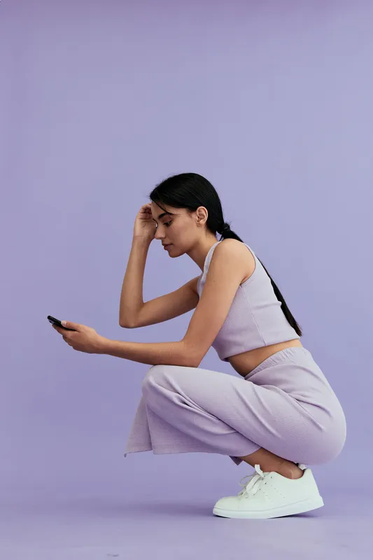 Young woman squatting and looking at smartphone with focused expression, wearing casual striped outfit and white sneakers on purple background.