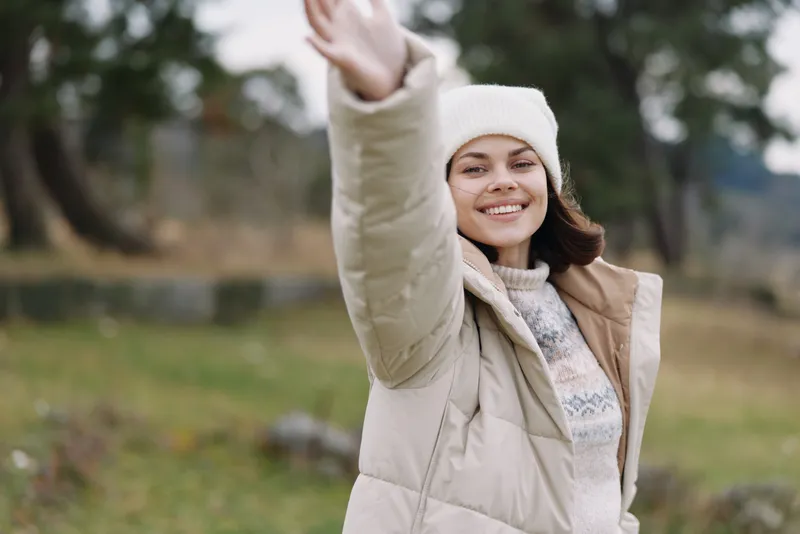 Woman outdoors waving smiling in a light winter coat and beanie, standing in an open park with soft natural light, casual fashion and a friendly greeting mood.