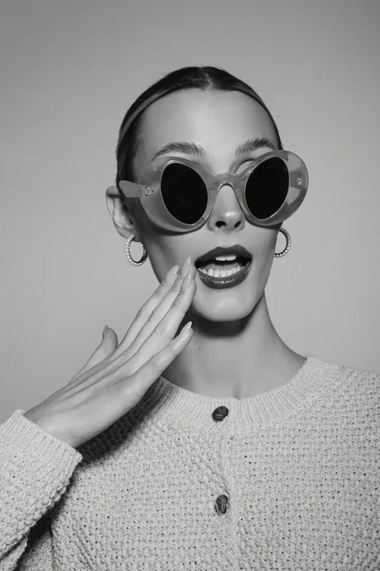 Stylish caucasian woman with light skin wearing round sunglasses and earrings expresses surprise and touches cheek in studio black and white portrait.