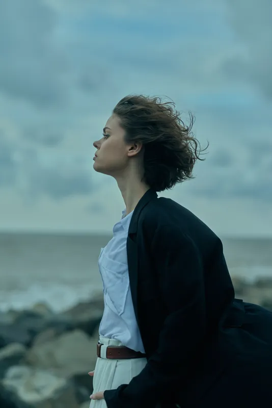 Profile of a windblown woman in a blazer by the sea, standing on the shore, facing the horizon with a calm, confident mood as the ocean breeze moves