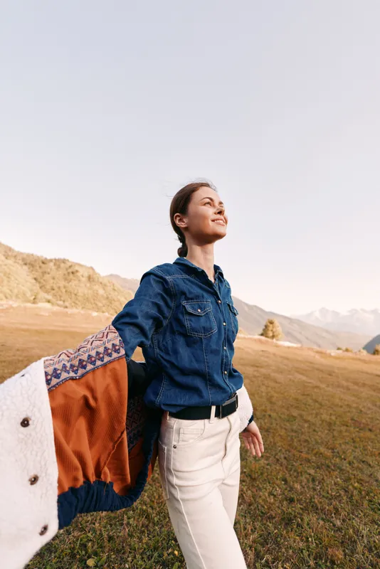 Woman holding jacket over shoulder in denim shirt and white pants, smiling in open field with mountains and clear sky, enjoying nature, freedom and a relaxed outdoor lifestyle.