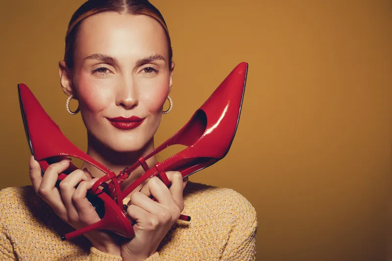 Smiling woman with red lipstick holds red high heel shoes near face on brown background. Concept of fashion, style, beauty, and confident female model portrait.