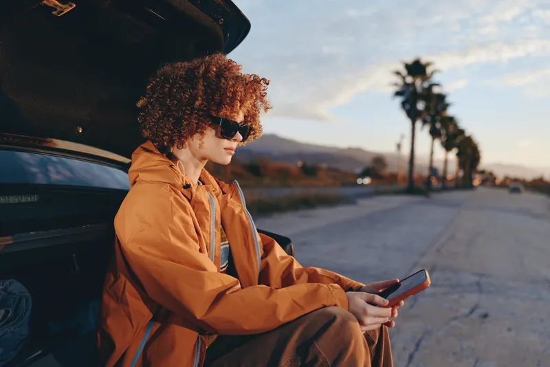 Woman lifestyle outdoors, orange jacket, leaning by car, using smartphone on road at sunset, curly hair, warm light, calm expression, palm trees, distant mountains, soft breeze
