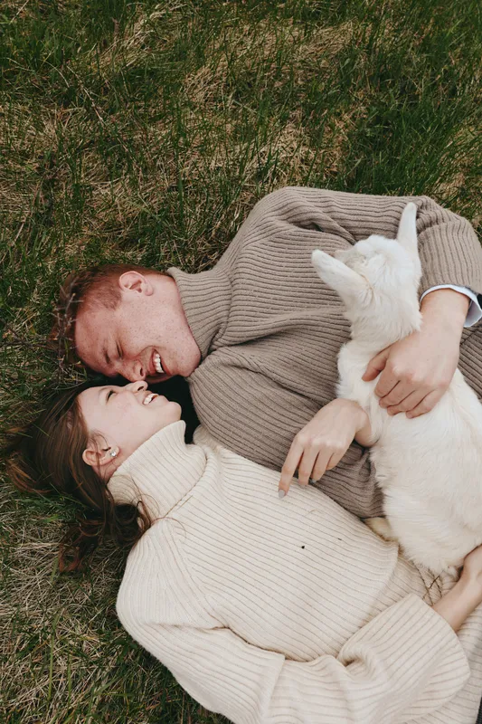 Happy couple relaxing on grass with a playful goat kid, embracing and smiling, casual knitwear, warm afternoon light, genuine affection, outdoors lifestyle moment