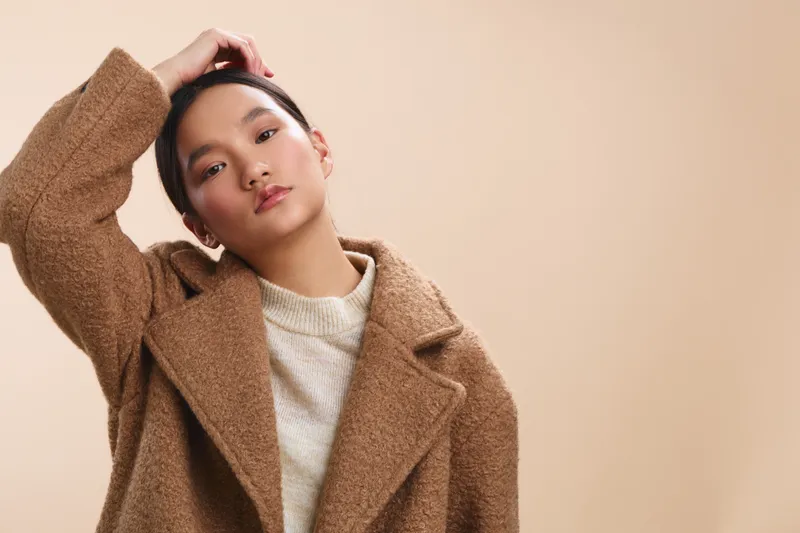 Asian woman wearing a cozy beige coat stands against a soft neutral backdrop, projecting a calm, stylish mood with a modern casual vibe and thoughtful gaze.