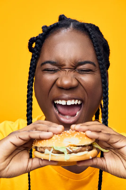Happy woman enjoying a burger against a vibrant yellow background, showcasing healthy eating habits and joy in food experiences