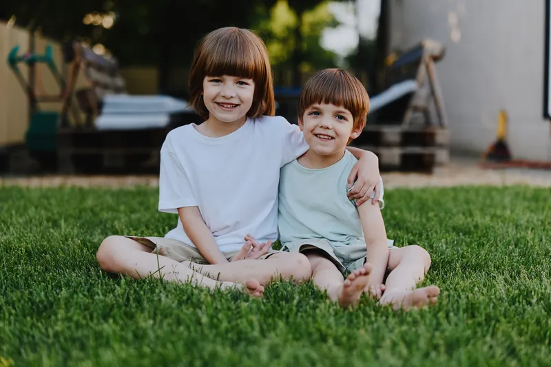 Happy children sitting on green grass, enjoying a sunny day together. Their smiles radiate joy and playfulness in a serene outdoor setting.