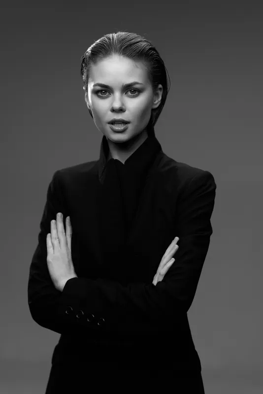 Black and white portrait of serious woman in dark jacket with crossed arms, monochrome, studio lighting, elegant and confident expression.